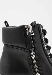Bottine en cuir noir avec une fermeture éclair argentée et un design à lacets, présentant une texture lisse et des détails de couture contrastés.