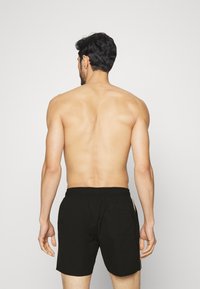 Shorts de bain noirs avec une taille élastique, une poche arrière et des bandes latérales, présentés sur un modèle vu de dos contre un fond uni.