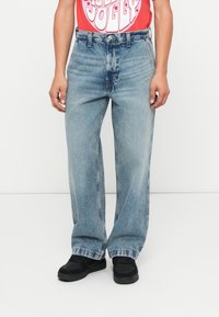 Svijetlo plave traperice slobodnog kroja, s visokim strukom i kontrastnim šavovima. Uparene s crnim cipelama i crvenom grafičkom majicom.