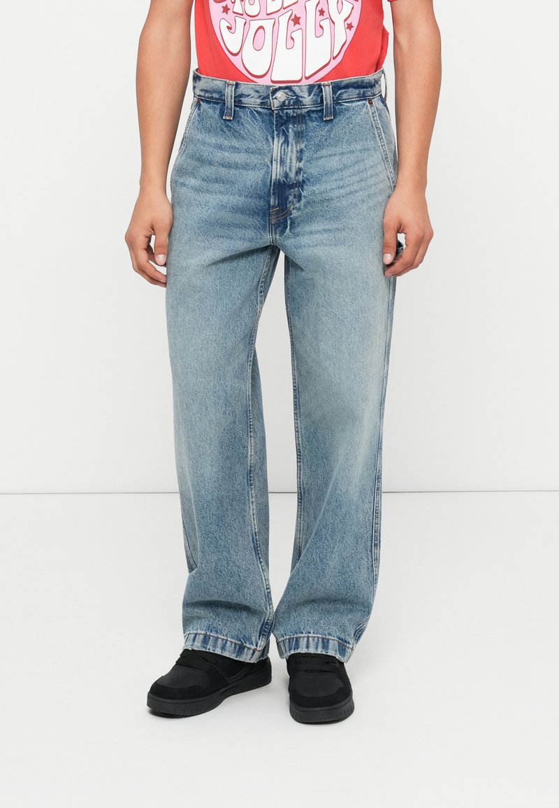 Světle modré džíny s volnějším střihem, s vysokým pasem a kontrastním prošitím. V kombinaci s černými botami a červeným tričkem s grafikou.