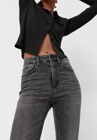 Personne portant un jean noir délavé taille haute et une chemise noire partiellement boutonnée, les bras levés, la section médiane visible.