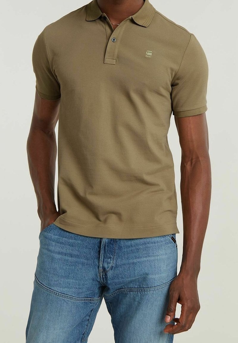 Homme portant un polo vert olive à manches courtes et un jean bleu, une main dans la poche, sur un fond uni.