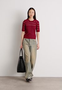 Burgunderfarbener Strickoberteil mit kurzen Ärmeln und Ausschnittdetails, kombiniert mit hellen, locker sitzenden Jeanshosen und einer schwarzen Tote-Tasche.