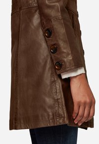 Brown leather jacket with a smooth texture, featuring large buttons along the side seam and a soft knit cuff peeking from the sleeve.