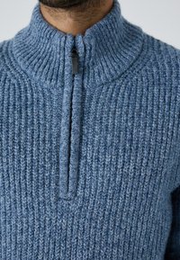 Zils adīts džemperis ar augstu apkakli, tajā ir pusfuča rāvējslēdzis priekšpusē. Audums ir ar teksturētu ribu rakstu, piedāvājot mājīgu izskatu.