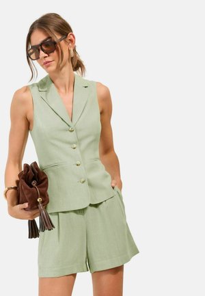 Žena oblečená v svetlozelenom bezrukávovom blejzri a šortkách, drží hnedú kabelku s strapcami, má na sebe slnečné okuliare a zlaté kruhové náušnice.