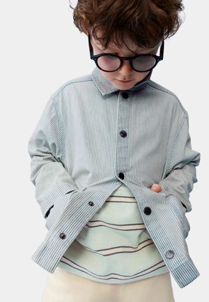 Bambino giovane con capelli ricci che indossa grandi occhiali rotondi, una camicia azzurra a righe con bottoni sopra una maglietta a righe, che guarda verso il basso con le mani nelle tasche.