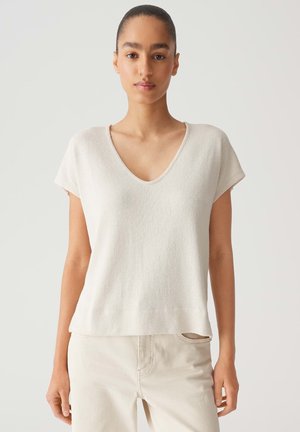 Frau mit kurzen Haaren, die ein hellbeiges kurzärmeliges V-Ausschnitt-Oberteil und helle Hose vor einem schlichten Hintergrund trägt.