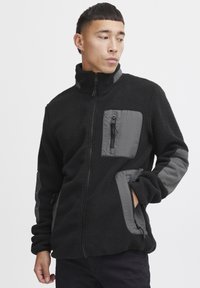 Svart fleecejakke med grå nylon detaljer, med høy krage, frontlommer med glidelås og en strukturert, myk overflate.