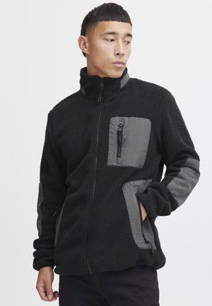 Svart fleecejakke med grå nylon detaljer, med høy krage, frontlommer med glidelås og en strukturert, myk overflate.