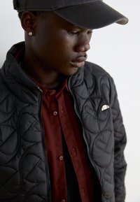 Melns ķīļveida jaku ar augstu apkakli, sudraba rāvējslēdzis, uzvilkts virs vīnogu krāsas pogājama krekla. Tam ir logo uz kreisā krūšu laukuma. Valkāta melna cepure.