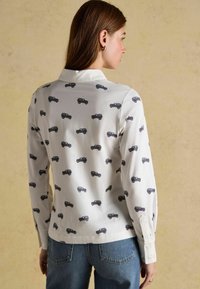 Biela košeľa s dlhým rukávom, ktorá má vzor malých čiernych áut. Hladká látka s golierom a gombíkovými manžetami. Spárovaná s modrými džínami.