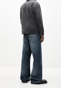 Persona vestida con una camiseta de manga larga gris oscuro, pantalones vaqueros de pierna ancha azules y zapatos negros, de pie frente a un fondo blanco liso.
