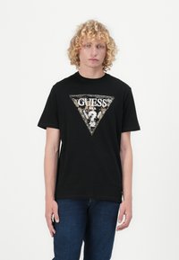 Svart bomull T-skjorte med et stort trekantet grafisk trykk med "GUESS" tekst, intrikate detaljer i gull og hvitt, korte ermer og rund hals.