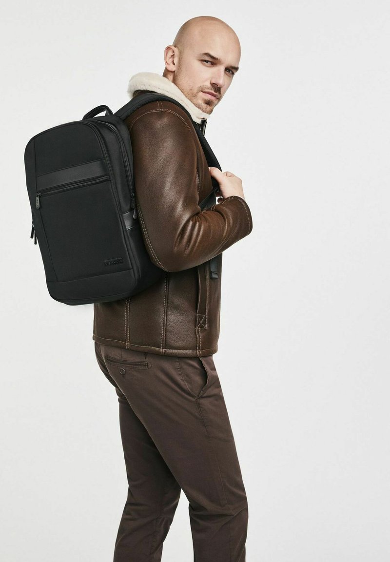 Mochila negra con respaldo acolchado, múltiples compartimentos con cremallera y un diseño texturizado, llevada sobre una chaqueta de cuero marrón.