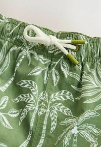 Costume da bagno verde con motivo di foglie di palma bianche, vita elastica con coulisse e occhielli in metallo per fissare la coulisse.