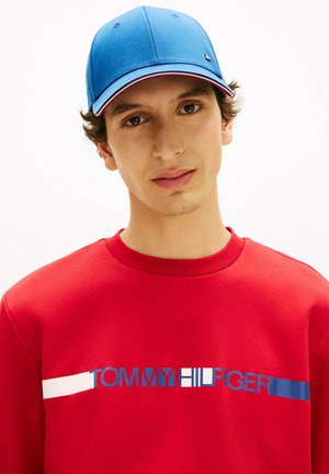 Giovane uomo con berretto blu e felpa rossa con la scritta e il logo "TOMMY HILFIGER" sul petto, su sfondo bianco.