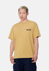 Camiseta de manga corta color mostaza con cuello redondo y un logo negro impreso en el lado izquierdo del pecho. Presenta un corte holgado y está hecha de material de algodón.