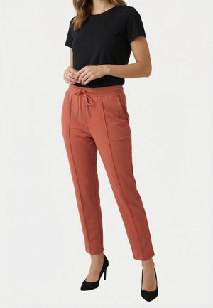 Mujer vistiendo pantalones de color óxido con cordón, camiseta negra de manga corta y tacones altos negros, posando contra un fondo blanco liso.