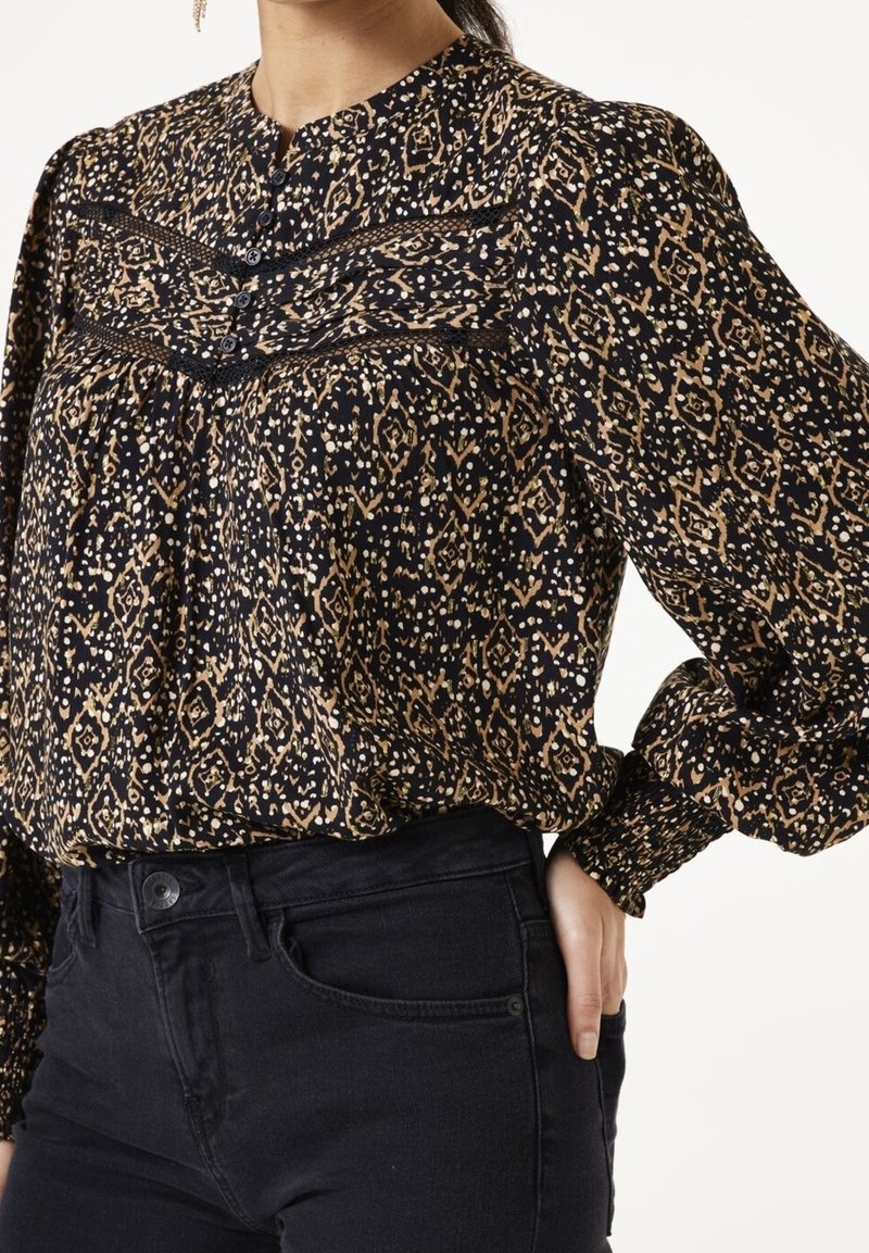 Femme portant un chemisier noir avec un imprimé géométrique beige, un yoke boutonné, des poignets froncés et un jean noir taille haute, main posée sur la hanche.