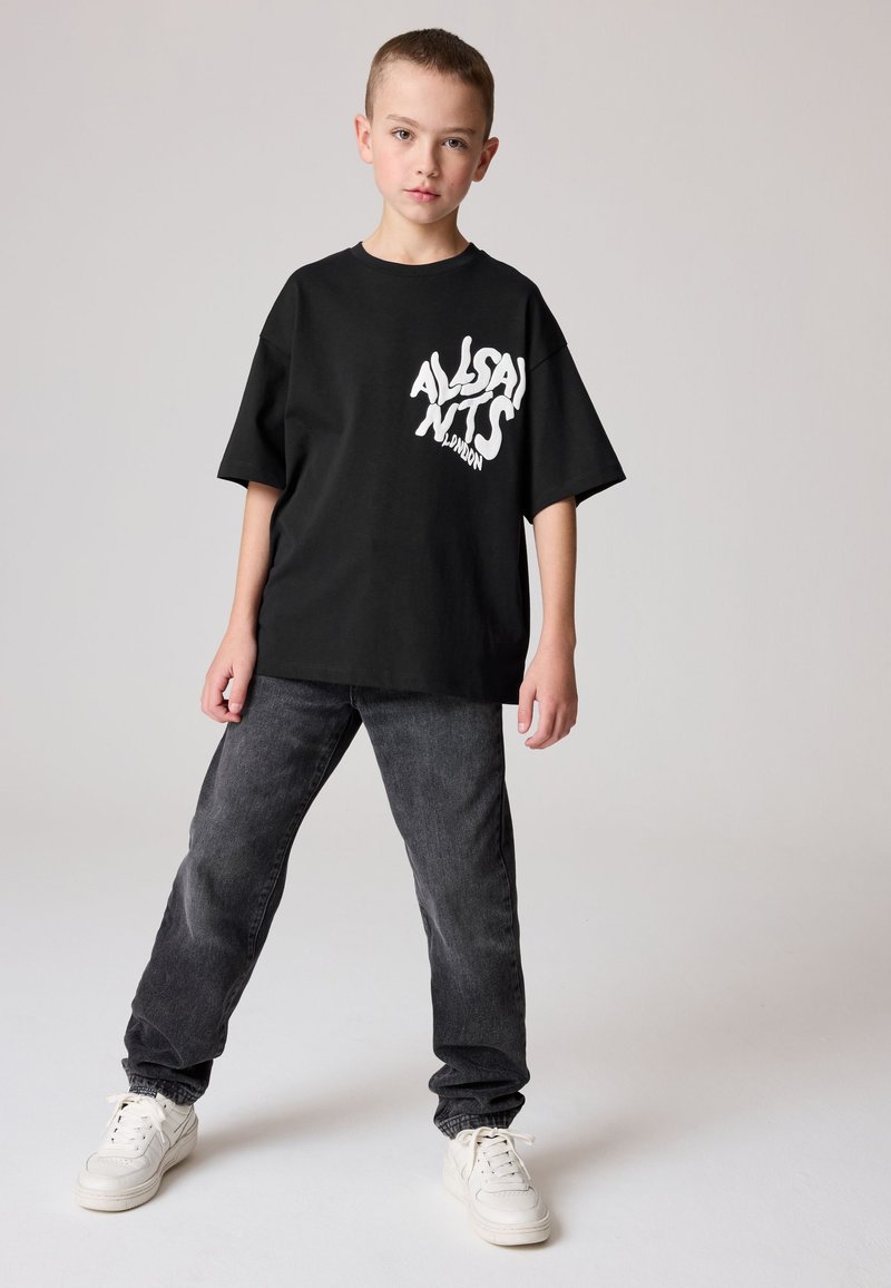 Junge trägt ein schwarzes oversized T-Shirt mit weißem Text, dunkle Jeans und weiße Sneaker, steht vor einem schlichten hellen Hintergrund.