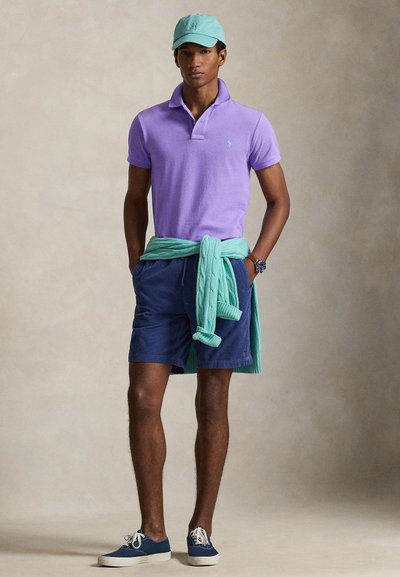 Mees kannab lavendlivärvi polo särki, türkiissinist nokamütsi, tumesiniseid lühkse, türkiissinist kampsunit, mis on seotud vöö ümber, ning tumesiniseid tossusid valgete paeltega, seisab käed taskutes.