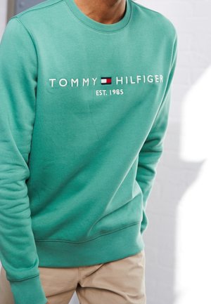Mężczyzna w morskim swetrze z napisem "Tommy Hilfiger EST. 1985" i logo, w połączeniu z beżowymi spodniami, stoi na jasnym tle.