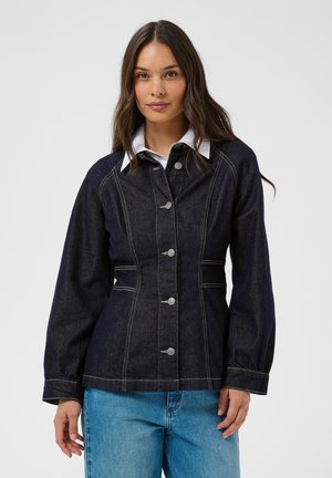 Femme aux longs cheveux foncés portant une veste en jean sombre boutonnée sur une chemise blanche à col et un jean bleu clair, debout devant un fond uni.