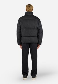 Černá prošívaná bunda, prošívaná textura, vysoký límec, nošená přes černé kalhoty a boty, ukazující uvolněný střih a minimalistický design.