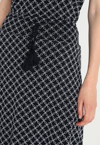 Tissu noir avec un motif géométrique blanc présentant des formes entrelacées. Comprend une ceinture tressée avec un détail de gland noir.