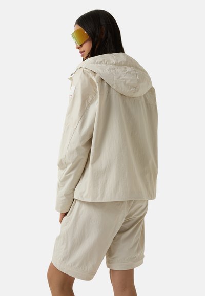 Beige letvægtsjakke med hætte, parret med matchende shorts. Stoffet har en blød tekstur og har minimal syning og detaljer.