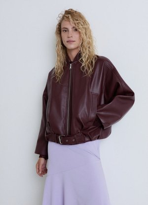 Persona con capelli biondi ricci che indossa una giacca di pelle rosso scuro con cerniera frontale e una gonna viola su uno sfondo chiaro uniforme.