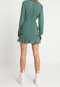 Vihreä kuvioitu mekko, jossa on röyhelöhelma ja pitkät hihat, istuva yläosa ja takana vetoketju. Paritettu valkoisten nilkkasaappaiden kanssa.