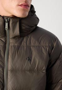 Nærbilde av en person som har på seg en mørkebrun puffjakke med høy krage og Calvin Klein "cK"-logoen på brystet.