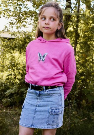 Pige iført en lyserød hættetrøje med sommerfuglegrafik og et denimskørt står udendørs med hænderne bag ryggen og ser til siden.