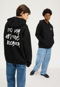 Two young people wearing black hoodies and loose blue jeans, one facing forward, the other showing hoodie's back with white text.