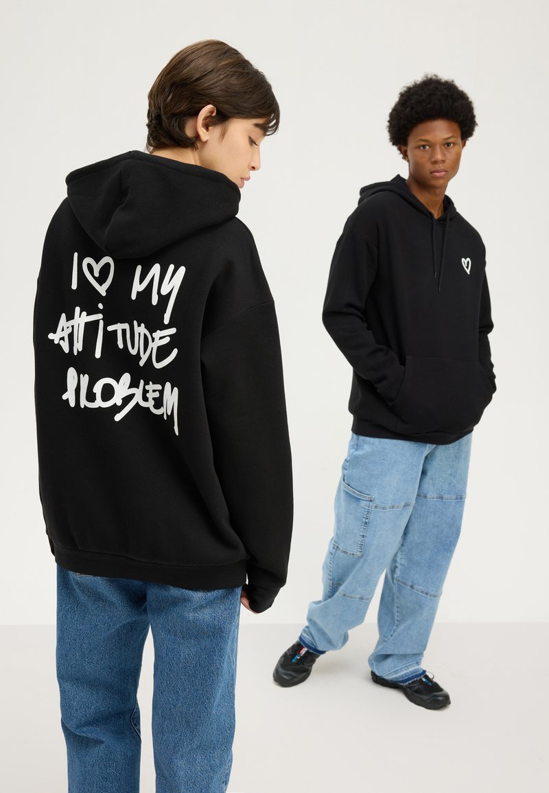 Two young people wearing black hoodies and loose blue jeans, one facing forward, the other showing hoodie's back with white text.