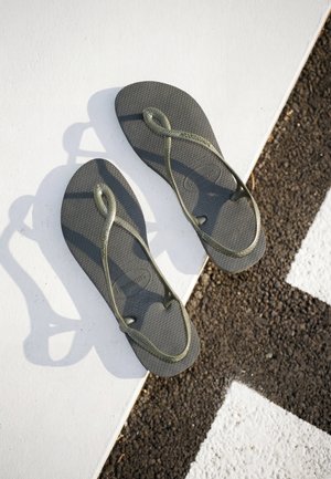 Sandalias Havaianas grises con tiras en lazos colocadas sobre una superficie blanca junto a un pavimento texturizado en blanco y negro.