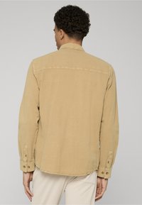 Chemise beige à manches longues en tissu doux, avec un col pointu, des poignets boutonnés et un ourlet droit.