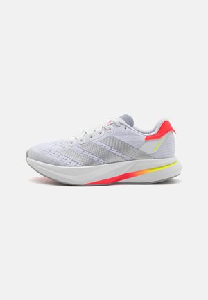 Zapatilla deportiva blanca con parte superior de malla, acentos grises, rayas reflectantes y suela en degradado de naranja brillante a amarillo. Cuenta con una entresuela acolchada.