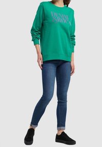 Sudadera verde con gráfico "DENIM" en plateado, combinada con jeans azules ajustados y zapatillas negras, mostrada contra un fondo neutro.