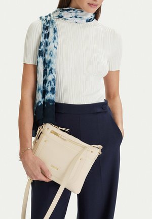 Mujer con camiseta blanca de manga corta acanalada y pantalones azul marino, bufanda azul con estampado, sujetando un bolso beige cruzado con detalles dorados.