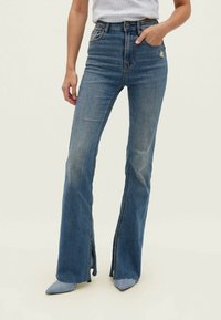 Vaqueros acampanados de acabado suave en denim azul medio, con una cintura alta, aberturas laterales en el dobladillo y un sutil desgaste en la tela.