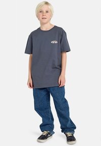 Blond dítě stojí, má na sobě volné tmavě šedé tričko, modré džíny a černé tenisky s bílými tkaničkami, na jednoduchém pozadí.