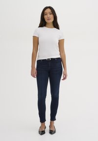 Ženska z dolgimi temnimi lasmi, oblečena v belo kratko majico s kratkimi rokavi, temno modre skinny kavbojke z zadrgama pri gležnjih in črnimi koničastimi petami.
