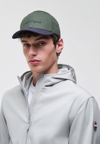 Fiatal férfi rövid, göndör hajjal, zöld és fekete sapkában, világosszürke cipzáras kapucnis dzsekiben, egyszínű háttér előtt.