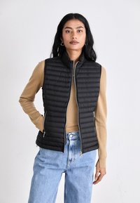 Črn, prešit brezrokavnik z visokim ovratnikom in zadrgo, oblečen čez bež pulover. Brezrokavnik ima dva žepa z zadrgo in prilegajočo silhueto.