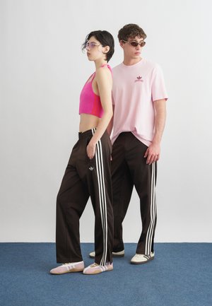 Δύο μοντέλα φορούν καφέ αθλητικό παντελόνι Adidas με λευκές ρίγες· το ένα με ροζ crop top και παπούτσια μπαλέτου, το άλλο με ανοιχτό ροζ T-shirt και αθλητικά παπούτσια.