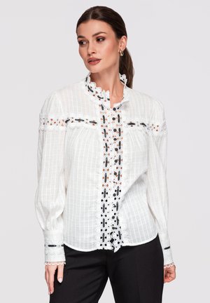 Femme portant un chemisier en dentelle blanche avec des détails en ruban noir, manches longues bouffantes, et un pantalon noir, regardant sur le côté sur un fond neutre.