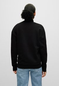 Schwarzer Pullover mit hohem Kragen, abgesenkten Schultern und gerippten Bündchen, kombiniert mit hellblauen Jeans. Besitzt eine glatte Textur.